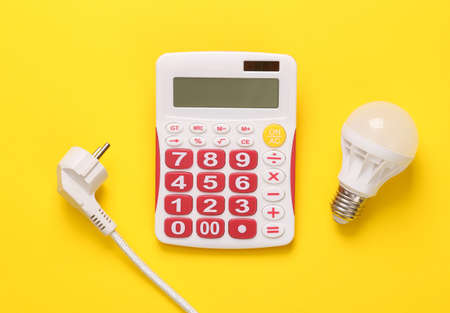 Calculation of the cost of electricity. Calculator, electric plug and energy-saving light bulb on a yellow background. Top view. flat layの写真素材