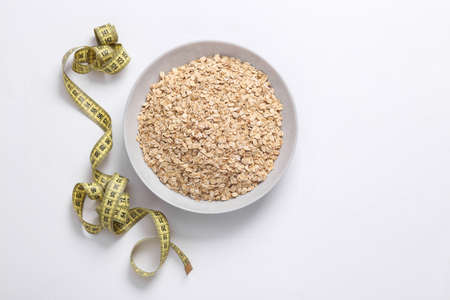 Bowl of raw oatmeal with a measuring tape on a white background. Diet, weight loss concept