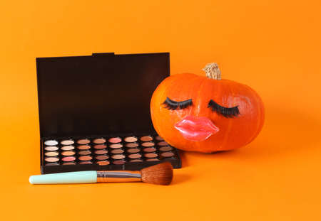 Pumpkin with false eyelashes and lips, eye shadow palette and makeup brush on orange background. Minmal beauty concept, creative layoutの写真素材
