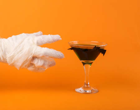 Mummy's hand wrapped in white bandages reaches for a glass of cocktail on an orange background. Halloween party concept.の写真素材