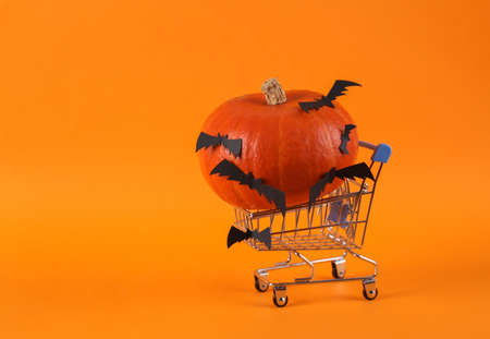 Shopping trolley and pumpkin with bats on orange background. Minimal halloween still life, decor, creative layout.
