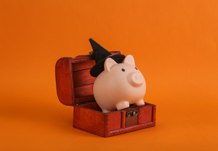 Halloween still life. Piggy banks with a witch's hat in a chest, orange backgroundの写真素材