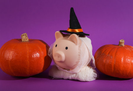 A piggy bank with a witch's hat and wrapped in bandages and pumpkins on a purple background. Trick or treat. Halloween still lifeの写真素材