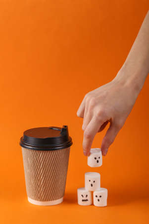 Hand takes halloween marshmallow ghost and coffee cup on orange background.の写真素材