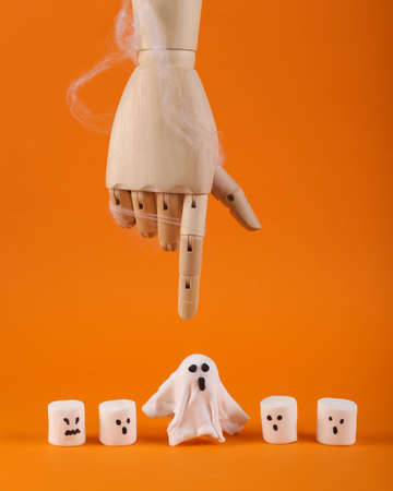 Halloween still life. Wooden puppet hand points to a ghost among halloween marshmallows with facesの写真素材