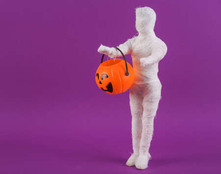 Trick or treat. Mummy doll wrapped in bandages with a jack-o'-lantern candy bucket on a purple background. Halloween minimal still life. creative layoutの写真素材