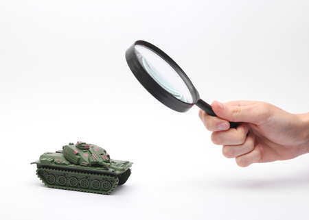 Hand holds magnifying glass and looks through and examines a plastic toy tank on a white background
