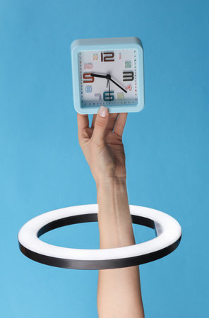 Woman's hand holds clock through led ring lamp on blue background. creative ideas.