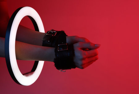 Woman's hands in sex shop leather handcuffs through led ring lamp, dark red neon light. sex games. creative ideas.の写真素材