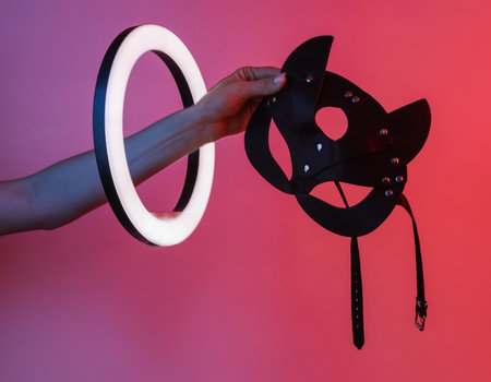 Woman's hand holds sex shop leather mask through led ring lamp in neon gradient light. sex games....