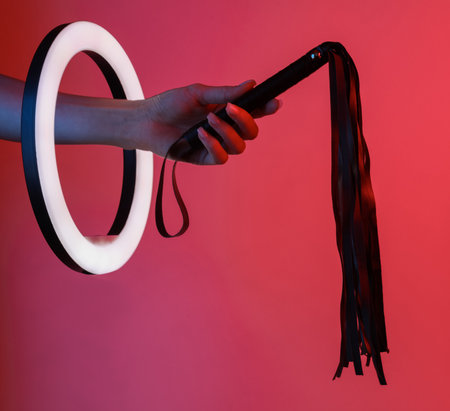 Woman's hand holds sex shop leather whip through led ring lamp in neon light. sex games. creative ideas.の写真素材
