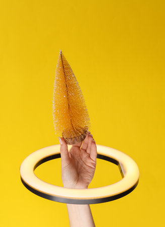 Woman's hand holds mini christmas tree through led ring lamp on yellow background. creative ideas.の写真素材