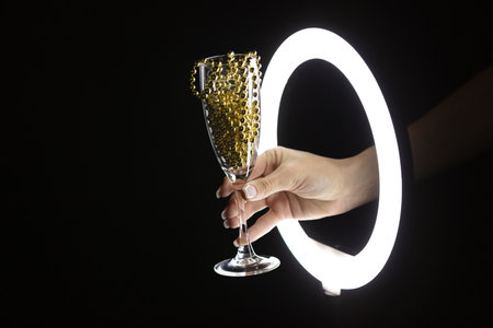 Female hand holding glass with garland through led ring lamp on black background. christmas party...
