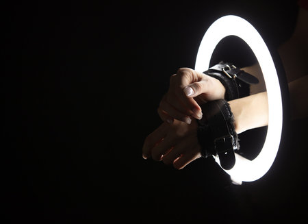Female hands in a sex shop handcuffed through led ring lamp on black background. sex concept
