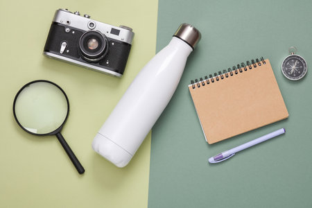 Hiking equipment. Thermos bottle, camera, magnifier, notebook and compass on green background. top...