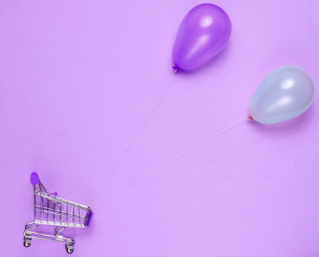 shopping concept. Mini shopping trolley, blue and pink balloons on purple background. Top view, minimalismの写真素材