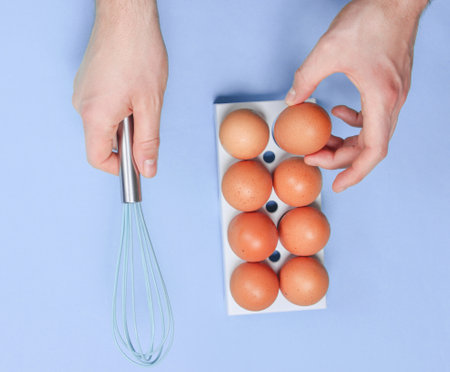 Women's hand taking hen's egg from eggs tray and whisk on blue background. Culinary concept, top view, flat lay, minimalismの写真素材