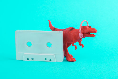 Toy red dinosaur tyrannosaurus rex with headphones and audio cassette on a turquoise background. Minimalism creative layout. Music lover. Retro 80sの写真素材