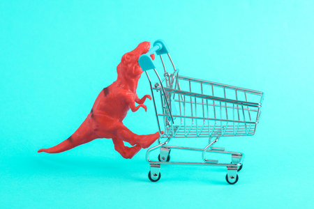 Toy red dinosaur tyrannosaurus rex with supermarket trolley on a turquoise background. minimalism creative layoutの写真素材