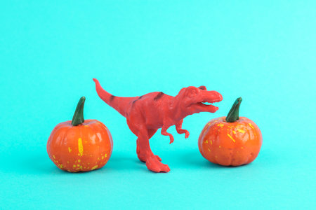 Toy red dinosaur tyrannosaurus rex with halloween pumpkins on a turquoise background. Minimalism creative layout. Trick or treatの写真素材