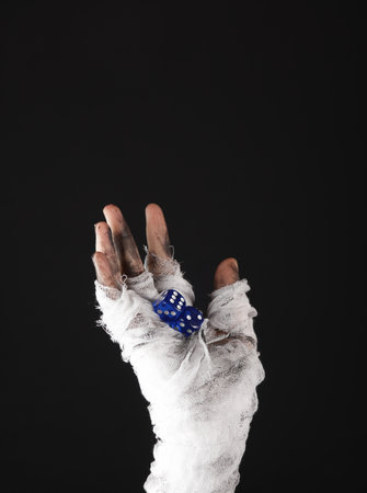 Mummy's hand wrapped in bandage holds dice isolated on black background. halloween conceptの写真素材