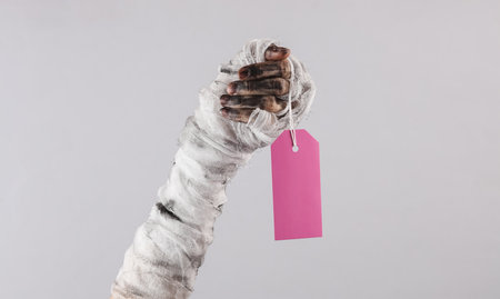 Mummy hand holding empty price tag isolated on gray background. Halloween sale conceptの写真素材