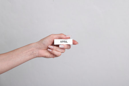 Woman's hand holds wooden white plate with inscription april on a gray backgroundの写真素材