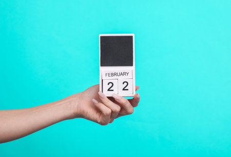 Female hand holding wooden calendar with date February 22 on blue backgroundの写真素材