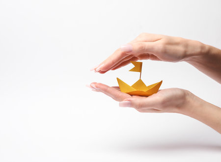 Hands holding and protecting yellow paper boat on white background. Concept of protection, insurance, securityの写真素材