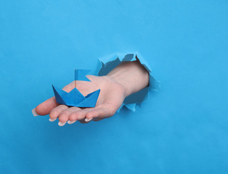 Female hand holds paper boat through torn hole in blue paperの写真素材