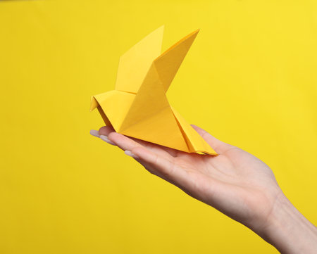 Female hand holding origami paper dove on yellow backgroundの写真素材