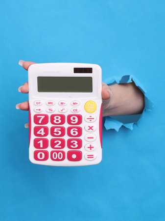 Female hand holds calculator through torn hole of blue paperの写真素材