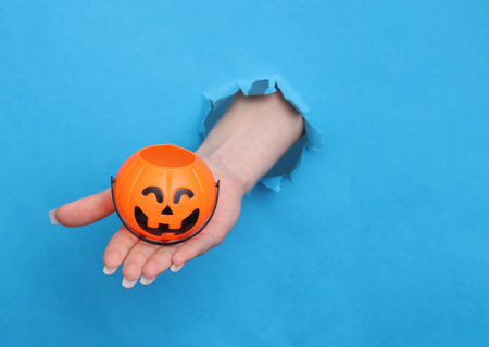 Female hand holds Halloween basket through torn hole of blue paper. Trick or Treatの写真素材