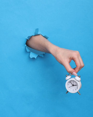 Female hand holds alarm clock through torn hole of blue paper