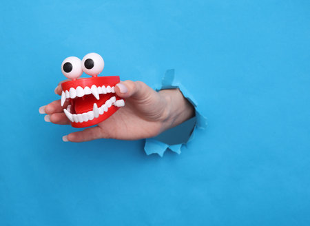 Female hand holds toy joke jaw through torn hole of blue paperの写真素材