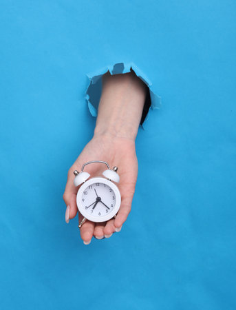 Female hand holds alarm clock through torn hole of blue paperの写真素材