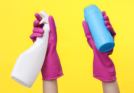 Hands in purple rubber cleaning gloves holding detergent bottles on a yellow background. House cleaning and housekeeping conceptの写真素材
