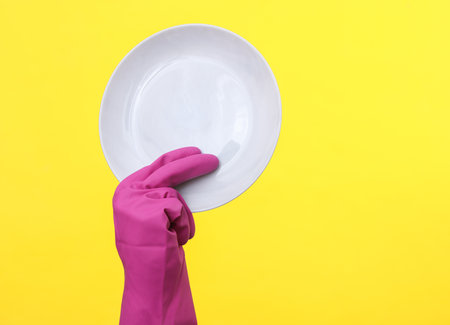Hand in purple rubber cleaning glove holding plate on a yellow backgroundの写真素材