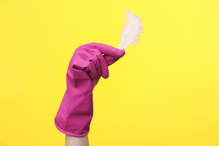 Hand in purple rubber cleaning glove holding feather on a yellow background. House cleaning and housekeeping conceptの写真素材