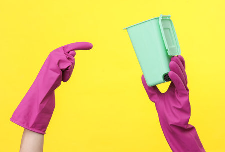 Hands in purple rubber cleaning gloves with mini trash can on a yellow background.の写真素材