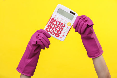 Hands in purple rubber cleaning gloves holding calculator on a yellow background. House cleaning and housekeeping conceptの写真素材