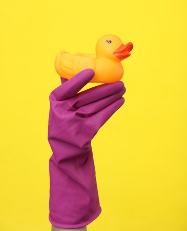 Hand in purple rubber cleaning glove holding rubber duck on a yellow background. House cleaning and...