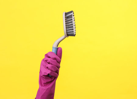Hand in purple rubber cleaning glove holding brush on a yellow background. House cleaning and housekeeping conceptの写真素材
