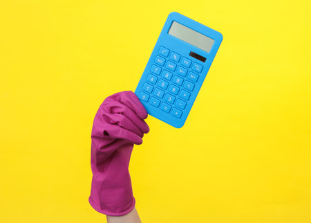 Hand in purple rubber cleaning glove holding calculator on a yellow background. House cleaning and housekeeping conceptの写真素材