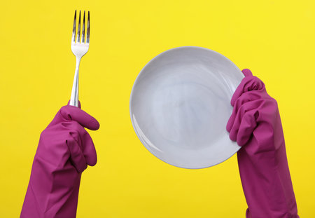 Hands in purple rubber cleaning gloves holding plate and fork on a yellow backgroundの写真素材