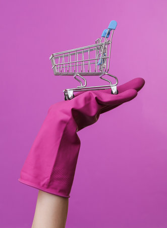 Hand in purple rubber cleaning glove holding mini supermarket trolley on a purple background. House cleaning and housekeeping, shopping conceptの写真素材
