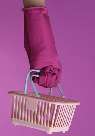 Hand in purple rubber cleaning glove holding mini supermarket basket on a purple background. House cleaning and housekeeping, shopping conceptの写真素材