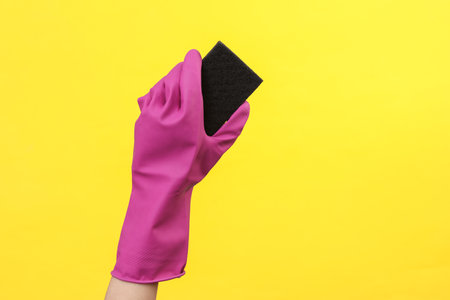 Hand in purple rubber cleaning glove holding sponge on yellow background. House cleaning and housekeeping conceptの写真素材