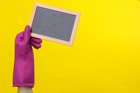 Hand in purple rubber cleaning glove holding empty chalk board on a yellow background. House cleaning and housekeeping conceptの写真素材