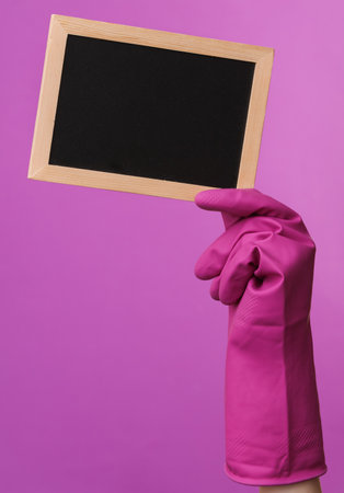 Hand in purple rubber cleaning glove holding empty chalk board on purple background. House cleaning...